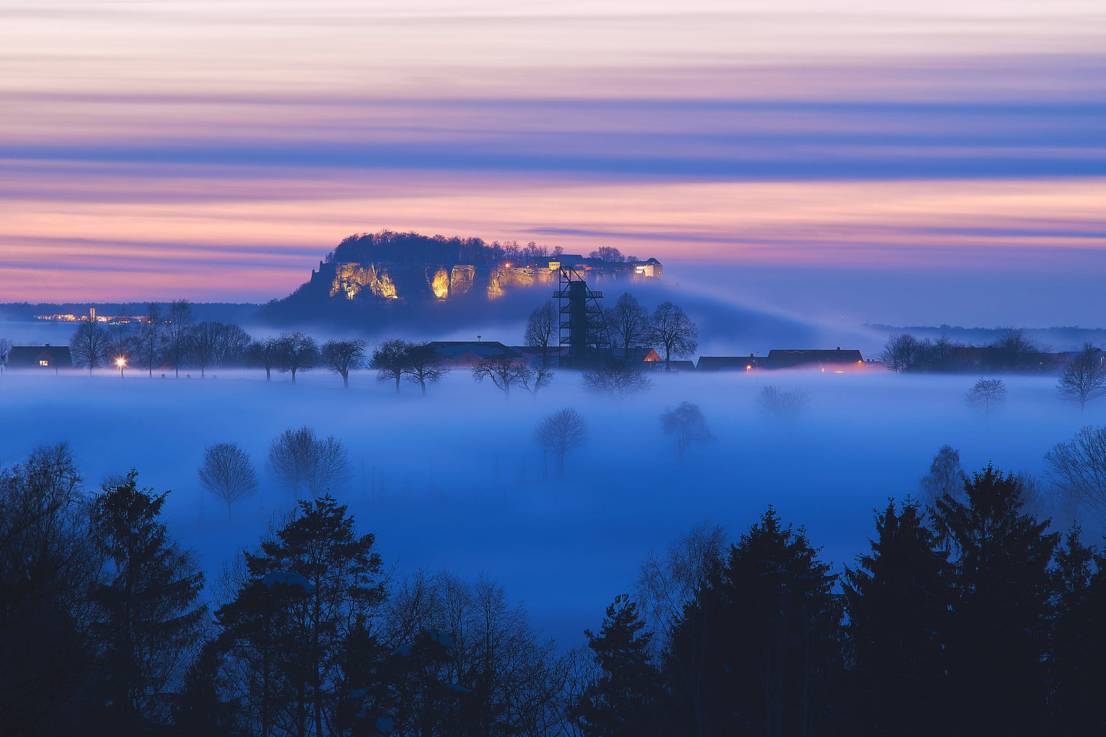 Die Festung Königstein nach Sonnenuntergang mit aufsteigendem Nebel. Davor sieht man den Aussichtsturm in Rathmannsdorf.